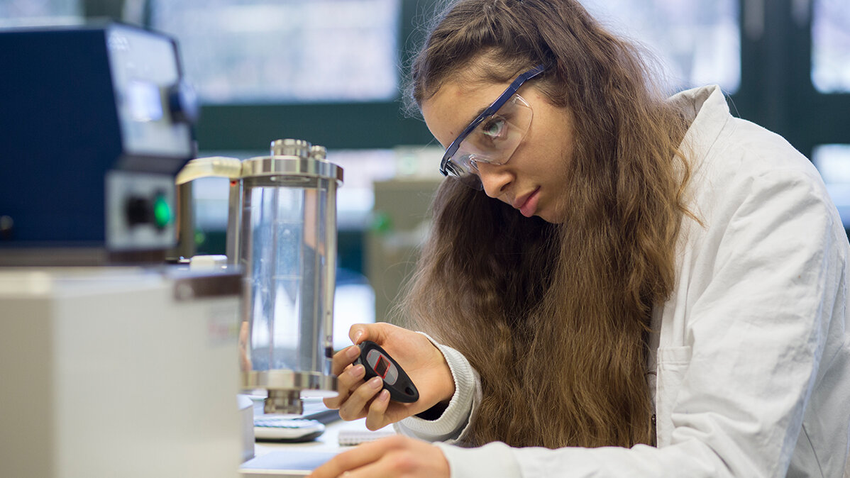 Eine junge Frau mit Schutzbrille untersucht mit einem Laborgerät eine Werkstoff-Probe.