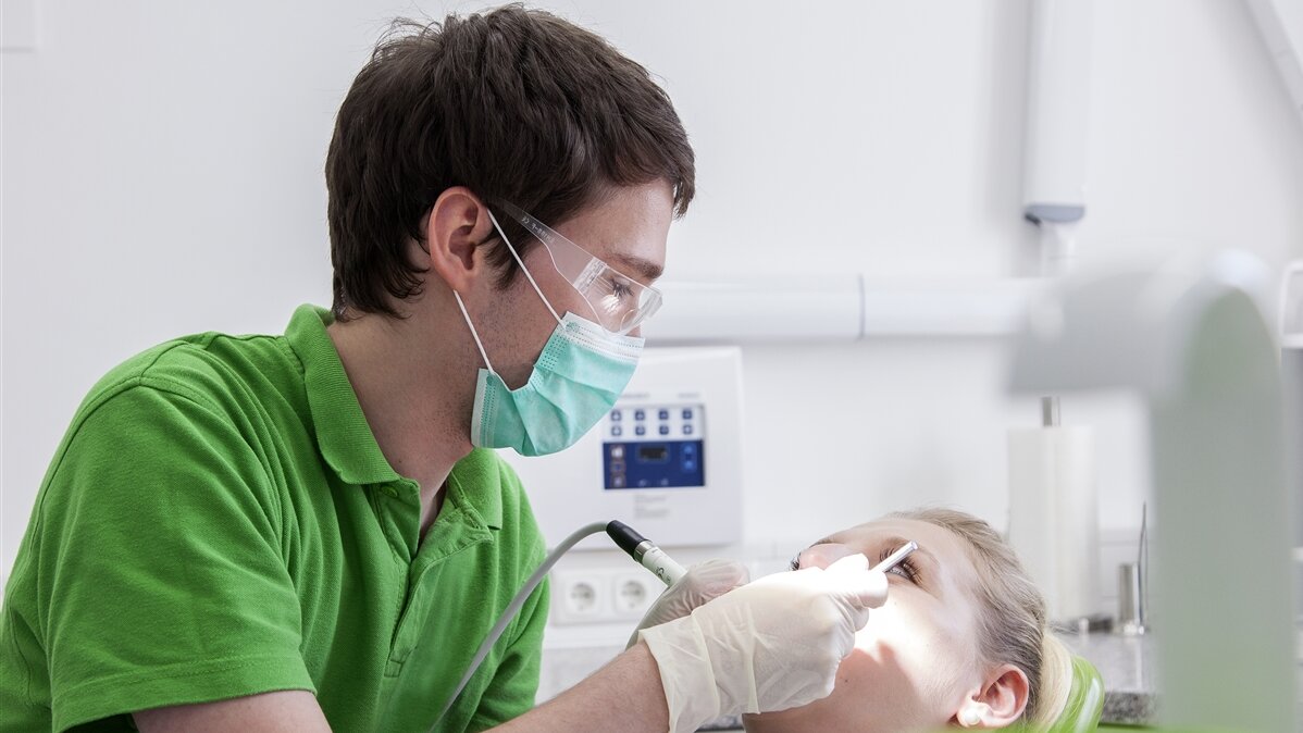 Ein junger Mann, mit Mundschutz, Latexhandschuhen und Schutzbrille ausgestattet, untersucht die Zähne einer junge Frau.