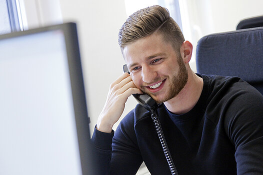Ein junger Mann sitzt vor einem Monitor in einem Büro und telefoniert. 