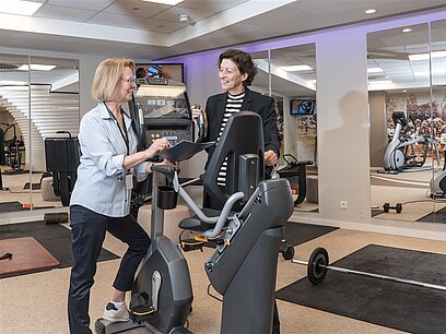 Eine Fitnessstudio-Mitarbeiterin steht mit einer Frau an einem Fahrradtrainer und erklärt den Trainingsplan.