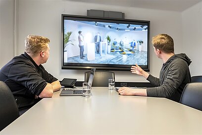 Zwei junge Männer besprechen ihre Idee mit großem Monitor und Laptop.