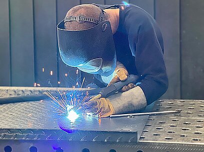 Ein Junger Mann in Schutzkleidung schweißt auf einer Werkbank Metallstücke zusammen.