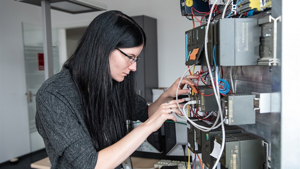 Eine junge Frau steht in einem EDV-Raum und prüft Kabel an einem Schaltkasten.