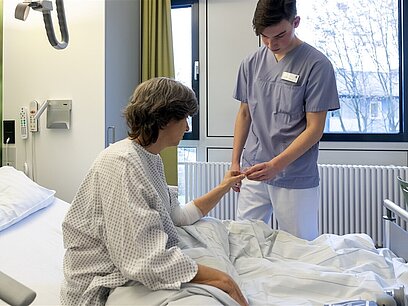 Ein junger Mann mit Pflegekittel gibt einer Frau im Krankenbett ein Medikament.