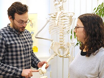 Ein Mann zeigt einer Frau einen Oberschenkelknochen an einem Modellskelett.