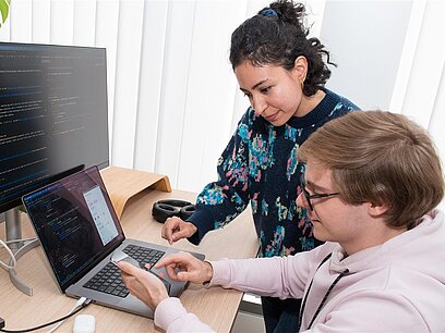 Eine Frau und ein junger Mann arbeiten mit einem Computer, einem Laptop und einem Handy.
