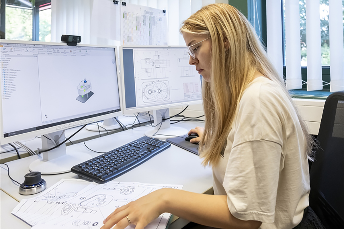 Eine junge Frau mit Beinem Bauplan entwirft am Computer ein 3-D Modell eines technischen Geräts.